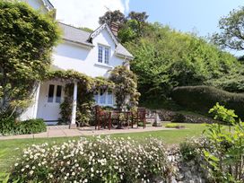 A white cottage with a tiled roof and outdoor wooden table with chairs on a patio surrounded by greenery and flowers at Lynch Cottage in Rousdon