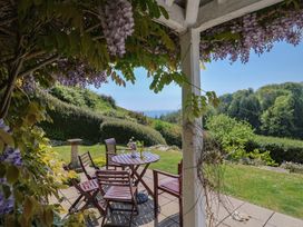 A patio with wooden chairs and a table under a pergola with hanging purple flowers at Lynch Cottage in Rousdon
