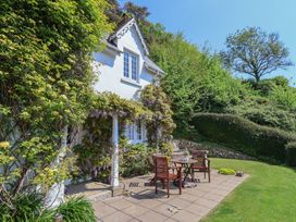 A patio with wooden chairs and a table outside a white cottage surrounded by greenery at Lynch Cottage in Rousdon