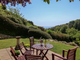 A garden patio with wooden chairs and table with glasses and bottle overlooking bushes and trees at Lynch Cottage in Rousdon
