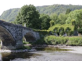 A stone bridge over a river with a cottage covered in greenery and a forested hill in the background at Mews Cottage in Llanrwst