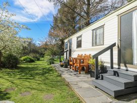An outdoor area with a white caravan on the right side a stone patio with wooden benches and potted plants and a grassy garden with trees and shrubs at Fron Dderw Caravan Llanfairynghornwy near Llanfechell