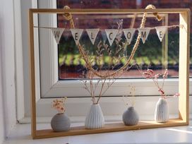 A window display with a wooden frame holding a welcome banner and four small vases with dried flowers at Banks View Filey in Filey