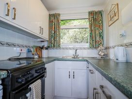 A kitchen with a stove, sink, kettle, cabinets, utensil holder, patterned curtains, and a wall sign at 8 The Park in Kilkhampton