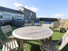 An outdoor patio area with a round wooden table and chairs on grass near a stone building and a swimming pool at Penpethy Farm in Tintagel
