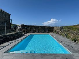 An outdoor swimming pool with lounge chairs along a stone wall and a house with patio furniture nearby at Penpethy Farm in Tintagel