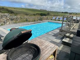 An outdoor swimming pool with blue water surrounded by a stone patio with a green grill and wooden table next to it and lounge chairs in the background at Penpethy Farm in Tintagel