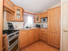 A kitchen with wooden cabinets, a stove, microwave, toaster, kettle, and a window with plaid curtains at Ty Newydd Caravan in Llannor near Pwllheli