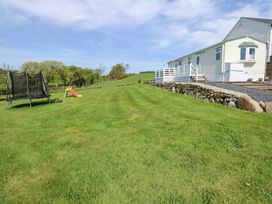 A large grassy yard with a trampoline and a slide next to a raised white caravan with a stone retaining wall at Ty Newydd Caravan in Llannor near Pwllheli