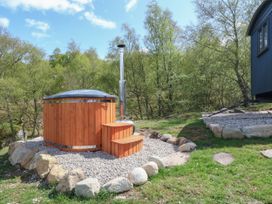 An outdoor wooden hot tub with steps and a metal chimney surrounded by gravel and stones near a blue cabin with trees in the background at Coraldie in Laggan near Newtonmore