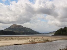 A sandy riverbank with water in the foreground and a forested hill under a cloudy sky at Coraldie in Laggan near Newtonmore