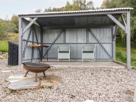 An outdoor shelter with two chairs and a table on a stone floor and a fire pit on gravel outside the shelter at Coraldie in Laggan near Newtonmore