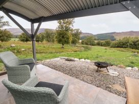 A covered patio with two wicker chairs and a fire pit overlooking a grassy field with trees and hills at Coraldie in Laggan near Newtonmore
