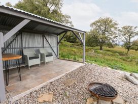 An outdoor seating area with two chairs under a metal shelter and a fire pit on gravel with grass and trees in the background at Coraldie in Laggan near Newtonmore