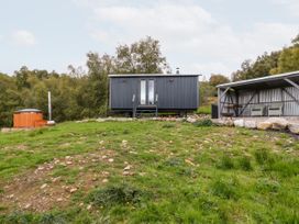 A small black cabin on wheels with stairs leading to the door a wooden hot tub and a covered seating area with two chairs in a grassy rural area at Coraldie Laggan near Newtonmore