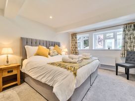 A bedroom with a grey upholstered bed with white bedding and yellow pillows beside wooden nightstands with lamps and a grey chair near windows with patterned curtains at Bedingfield House nr Debenham Debenham