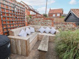 An outdoor seating area with wooden built-in benches, patterned cushions, a wooden bench, and a barbecue grill in a brick and stone walled garden at Dolphin Cottage Southwold