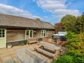 An outdoor patio with wooden bench table seating and a hot tub near a wooden building surrounded by greenery at Waldegrave Barn Hartest near Glemsford