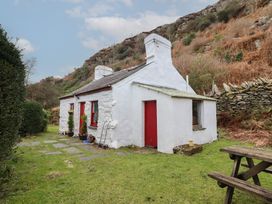 A small white stone cottage with red doors and windows in a grassy yard with a wooden picnic table and hillside behind at Quarry Cottage in Blaenau Ffestiniog