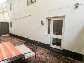 A narrow outdoor patio area with a metal bench and table covered by a striped cloth near a white building with a glass door and window at The White House in Torquay