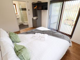 A bedroom with a double bed green pillows folded towels a gray blanket a wall-mounted TV an open wardrobe leading to a bathroom and vertical blinds on a glass door at Lodge 3 in Thorpe-On-The-Hill