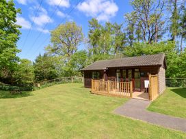 A small wooden cabin with a fenced porch on a grassy lawn surrounded by trees at Chalet Log Cabin L6 Combe Martin