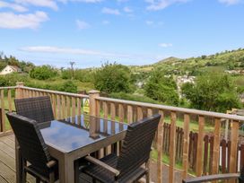An outdoor wooden deck with a glass table and four wicker chairs overlooking green hills and houses at Chalet Log Cabin L14 in Combe Martin