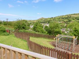 An outdoor fenced playground area with swings and a grassy field with hills and a village in the background at Chalet Log Cabin L14 Combe Martin