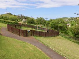 A small fenced playground with a tire swing and climbing frame in a grassy area with a paved path and hills in the background at Chalet Log Cabin L14 Combe Martin