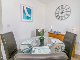 A dining area with a glass round table and four chairs with a flower vase a wall clock and a wooden sideboard at Ocean Retreat in Goodrington near Paignton