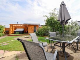 An outdoor patio with a glass table and four chairs with a closed umbrella a fire pit and a wooden shed with a hot tub in a garden at Branwen in Talacre