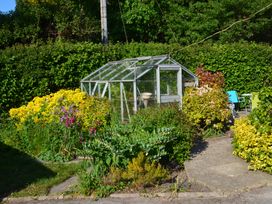 A garden with a small glass greenhouse surrounded by various plants and flowers with a seating area in the background