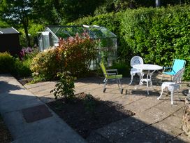 A patio area with a white metal table and chairs and a green and blue garden chairs near a greenhouse surrounded by plants at Woodstock in Chaffcombe near Chard