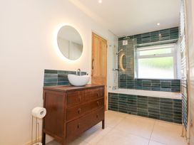 A bathroom with a wooden vanity with a vessel sink and round illuminated mirror and a bathtub with dark green tiles surrounding it at Ysgubor Maen Y Bardd in Rowen near Conwy