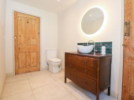 A bathroom with a wooden door a white toilet a wooden vanity with a white vessel sink a faucet a round illuminated mirror and soap dispenser at Ysgubor Maen Y Bardd Rowen near Conwy