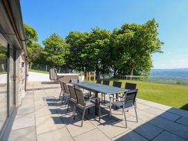 An outdoor patio with a glass table and twelve chairs a hot tub green lawn trees and a mountain view at Ysgubor Maen Y Bardd in Rowen near Conwy