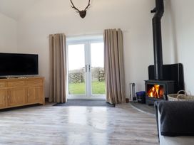 A living room with a wood stove a television on a wooden cabinet beige curtains and glass doors showing a garden at Gaerau Bach in Llanaber