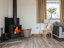 A wood burning stove with fire a basket and fire tools next to a window with curtains a lamp on a wooden side table and part of a sofa at Gaerau Bach in Llanaber