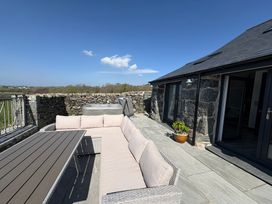 An outdoor patio with a sectional sofa and table next to a stone building and a stone wall with a hot tub behind Gaerau Bach in Llanaber
