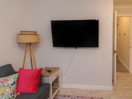 A living room with a wall-mounted television a floor lamp a side table with a remote control and a grey sofa with colorful cushions at 1 Ilsham Cottages in Torquay