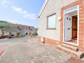 An exterior of a house with an open white door and a concrete pathway leading to the street with another house and trees in the background at Bryn Bach in Rhos-On-Sea