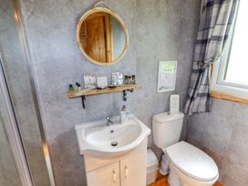 A bathroom with a white sink and cabinet a round mirror above a wooden shelf toiletries a white toilet a window with checkered curtains and a shower door at Moel Y Parc in Bryniau Clwyd near Denbigh