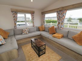 A living room with a grey L-shaped sofa with orange cushions and blankets a small wooden coffee table and patterned curtains at No30 Elm Rise in Gristhorpe near Filey