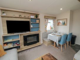 A living area with a television above a fireplace shelves with board games a dining table set for four with blue chairs and a window at No30 Elm Rise in Gristhorpe near Filey