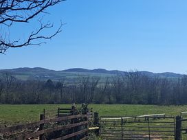 A grassy field with a metal gate and wooden fence with hills and trees in the background at Capel Park Tyddynod Bach in Llandyfrydog near Llanerchymedd