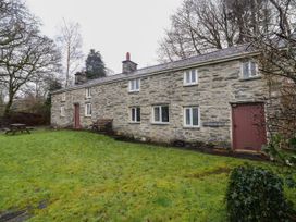 A stone cottage with a grassy yard bench and picnic table surrounded by trees at The Long House in Penmachno