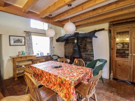 A dining room with a wooden table covered with a red floral tablecloth surrounded by wooden and wicker chairs with a fireplace and wooden beams at The Long House in Penmachno