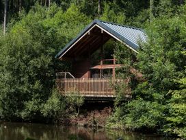 A wooden cabin with a deck surrounded by green trees by a pond at Deerpark Classic Golden Oak Waterside in Deerpark Forest