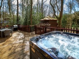 An outdoor wooden deck with a hot tub table and chairs and a barbecue grill surrounded by trees at Delamere Forest Golden Oak Treehouse in Delamere Forest