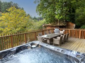 An outdoor deck with a hot tub dining table with chairs and barbecue grill surrounded by trees at Deerpark Classic Golden Oak Treehouse Deerpark Forest